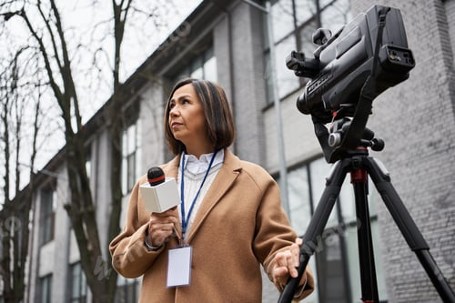 Preview: Multiracial female journalist reports live outside a building on a cloudy afternoon