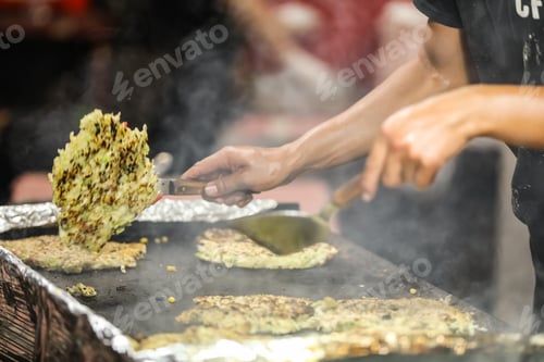 Preview: A Japanese man baking and flipping an Okonomiyaki on a grill outside at night.