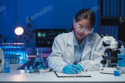 Preview: Young female scientist wearing lab coat and protective glasses writing notes on clipboard while work