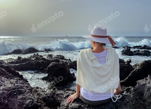 Preview: Back view of senior woman sitting on a rocky beach watching ocean waves crashing