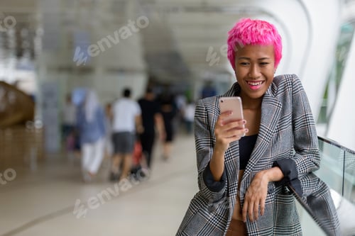 Preview: Happy young rebellious Asian businesswoman with pink hair using phone in the city