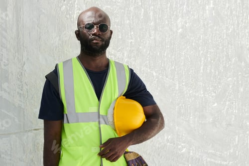 Preview: Serious black man in reflective vest and eyeglasses holding hardhat by his waist