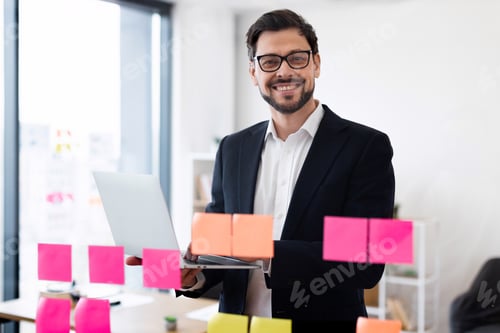 Preview: Smiling businessman working on laptop in modern office with sticky notes
