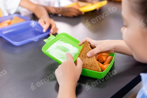 Preview: selective focus of schoolgirl taking sandwich from lunch box with fresh carrots near classmates