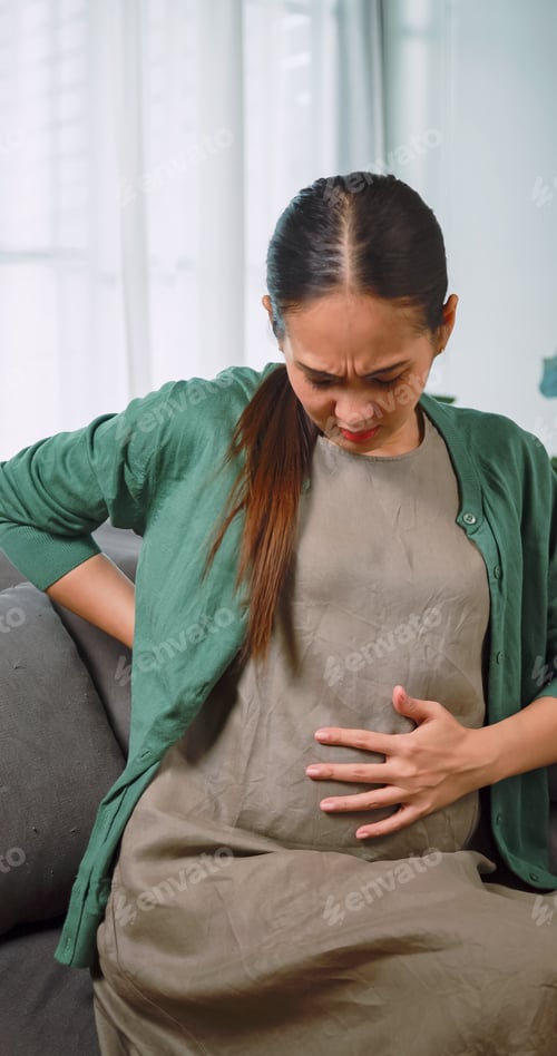 Preview: Asian pregnant woman having back pain while relaxing on sofa at home.