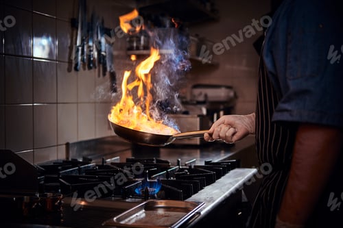 Preview: Chef wearing gloves and apron frying flambe on a pan in a dark restaurant kitchen