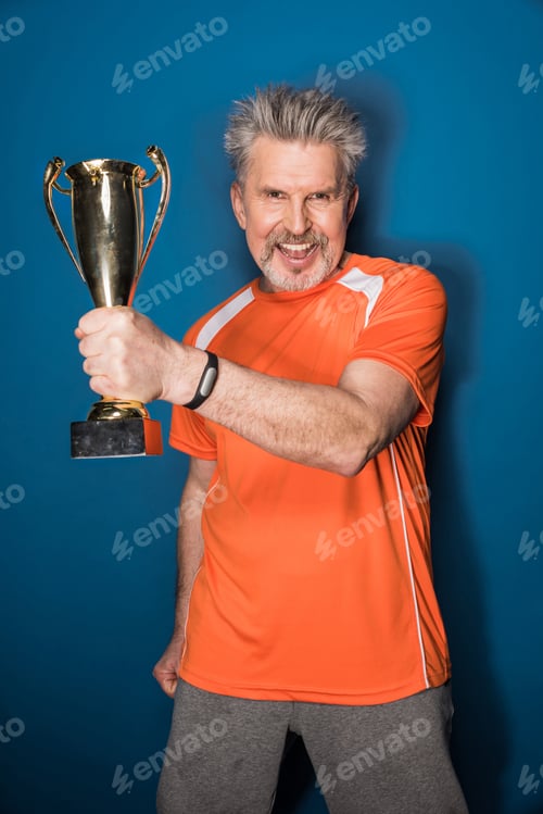 Preview: Excited mature sportsman holding trophy cup isolated on blue