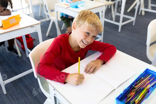 Visualização: Vista de alto ângulo de sorridente estudante do ensino fundamental caucasiano olhando para longe enquanto escrevia no livro na mesa