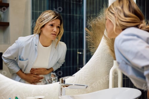 Preview: Mid adult woman suffering from stomach pain while standing in front of a mirror in the bathroom.