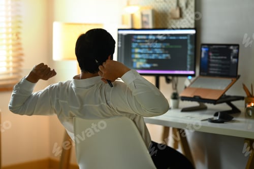 Preview: Back view of male programmer stretching arms and relaxing on chair at his workplace