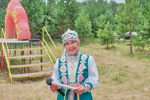 Preview: Asian mature woman in stylized Tatar clothes near stage. Concert host at village festival.