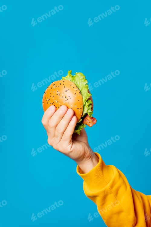 Preview: Male hand in yellow clothes hold hamburger on blue background