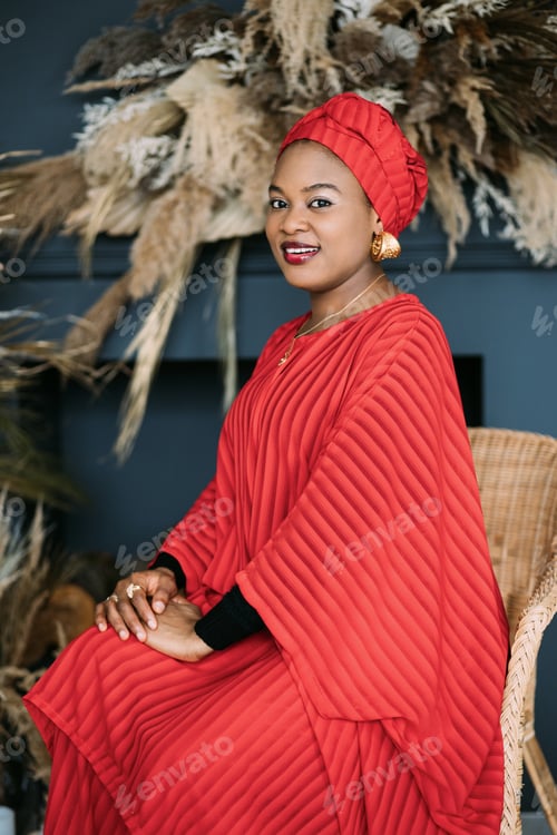Preview: Pretty African woman wearing an red fashionable suit and headscarf, sitting on the chair in studio