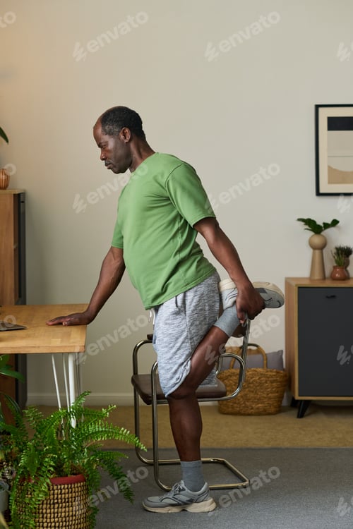 Preview: Senior Black Man Stretching Leg Indoors during Home Workout Routine