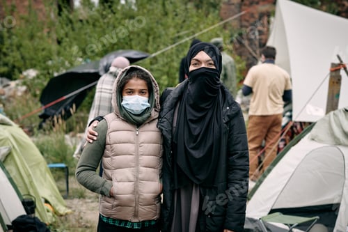 Preview: Young woman in hijab and her daughter standing against tent camp