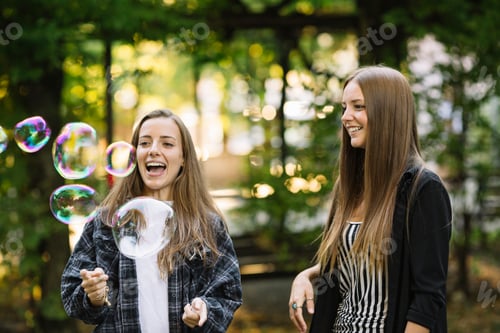 Preview: Two young female friends bursting floating bubbles in park