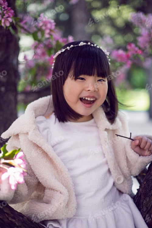 Preview: Korean girl in a white light fur coat and a headband sitting on a tree branch in a garden