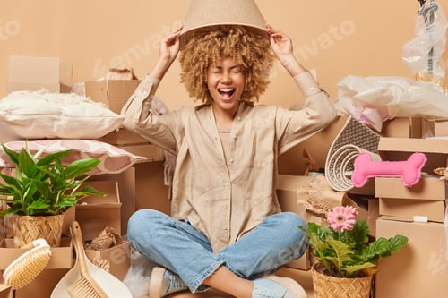 Preview: Cheerful young female renter sits crossed legs covers head with lampshade going to move and unpack