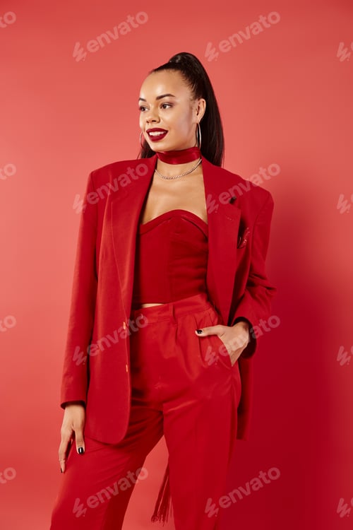 Preview: happy african american woman with ponytail posing with hand in pocket of pants suit on red backdrop
