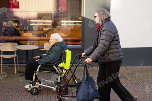 Vorschau: Reife Frau geht mit einer Gehhilfe neben einer älteren Frau im Rollstuhl vor dem Café