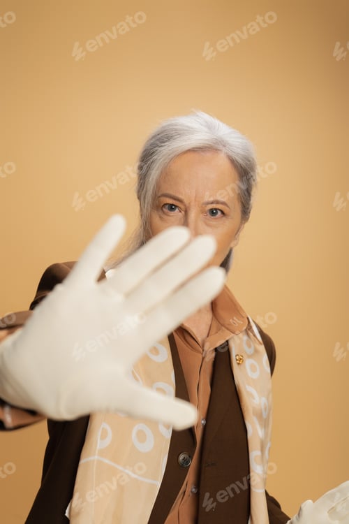 Preview: senior woman in white glove looking at camera while showing stop sign isolated on beige