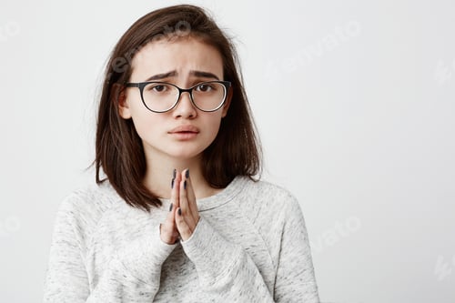Preview: Beautiful young dark-haired female in spectacles with begging look holding palms pressed together in