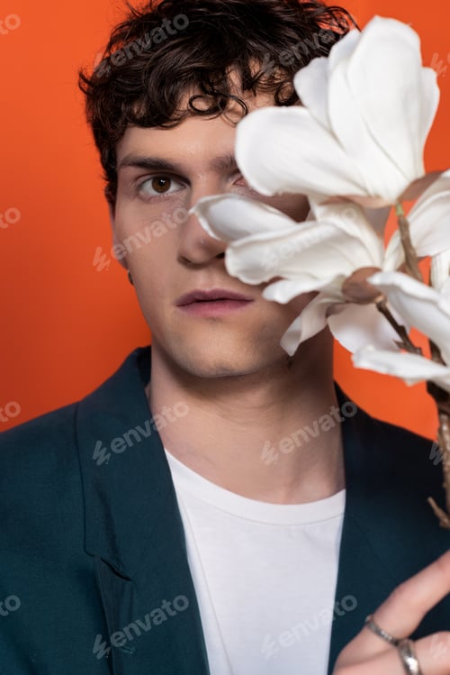 Preview: Curly man in jacket looking at camera near blurred magnolia flowers isolated on orange