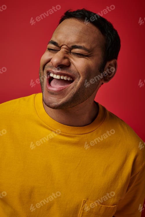 Preview: face expression, excited indian man in yellow t-shirt laughing on red background, open mouth