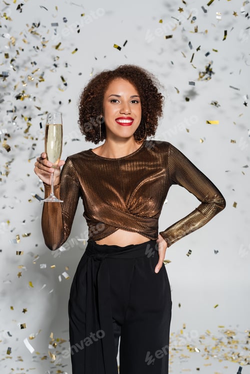 Preview: joyful african american woman standing with hand on hip and champagne glass near confetti on grey