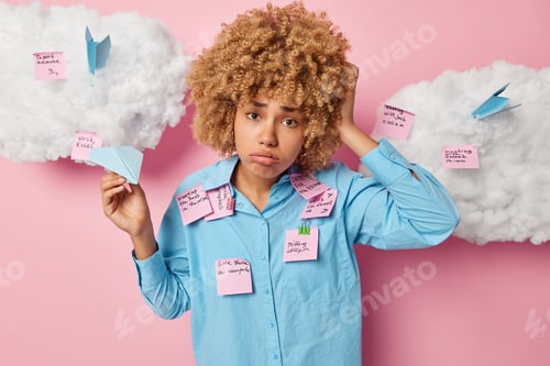 Preview: Upset curly haired female student holds paper made plane wears blue shirt covered with memo stickers