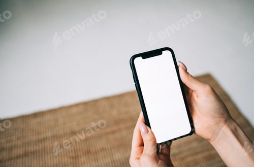 Preview: Woman holding a smartphone with a white screen mock up, in living room at home.