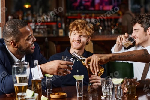 Preview: tattooed african american man pouring beer in glass of friend during bachelor party, interracial men
