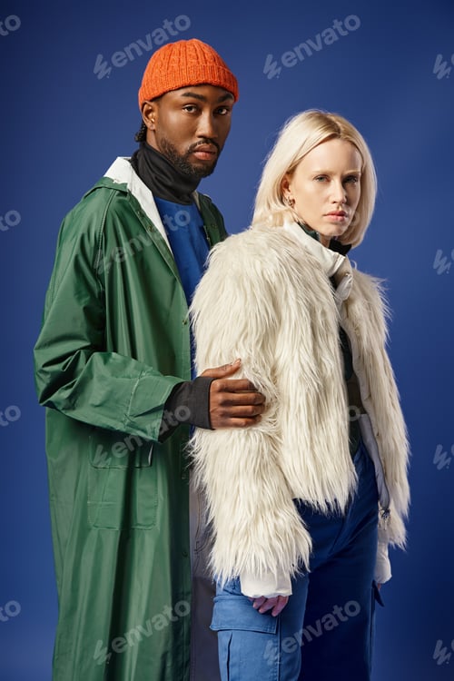 Preview: blonde woman posing with african american man in winter attire, stylish models on blue backdrop