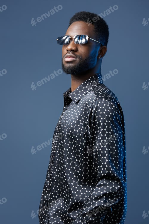 Preview: Stylish african man in sunglasses and polka dot shirt posing confidently against vibrant blue