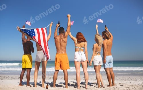 Preview: Rear view of diverse friends raising their hands on the beach while holding american flag