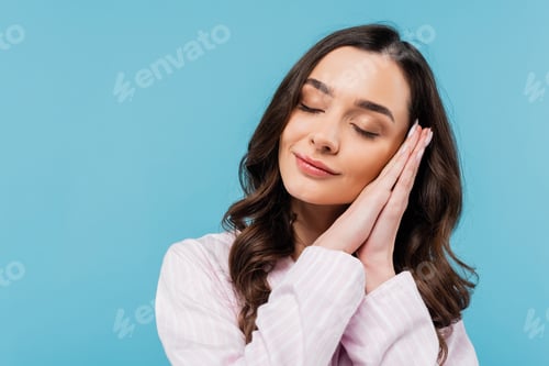 Preview: sleepy young woman with closed eyes folding hands while standing in pajamas isolated on blue