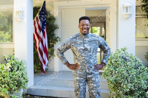 Preview: Happy african american male soldier with hands on hips outside home with usa flag, copy space
