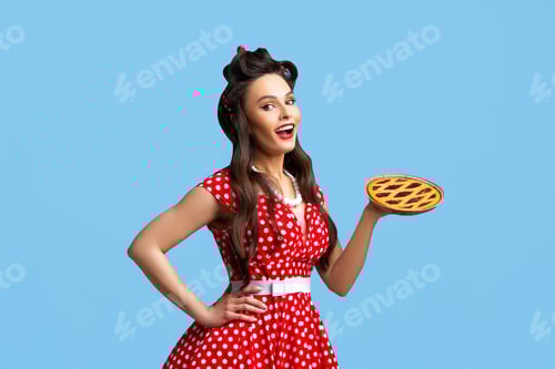 Preview: Happy pin up woman in polka dot red dress holding homemade sweet pie on blue studio background