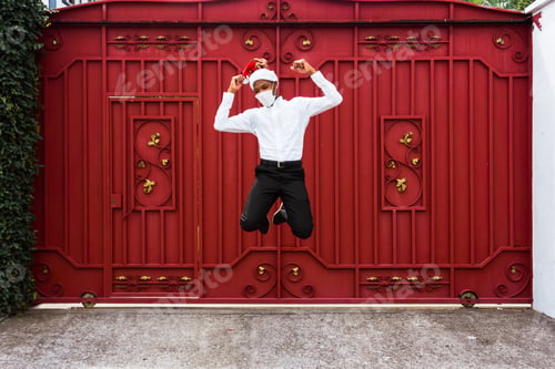 Preview: Young Afro-American man wearing a Santa hat and jumping in front of a red gate