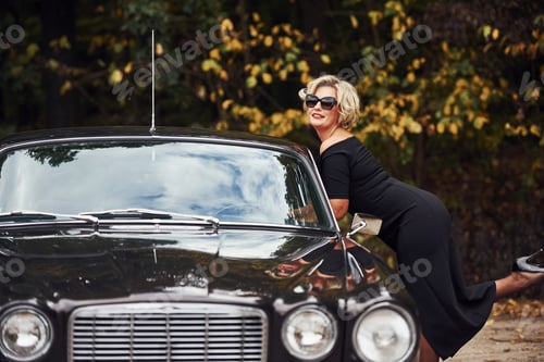 Preview: Blonde woman in sunglasses and in black dress near old vintage classic car