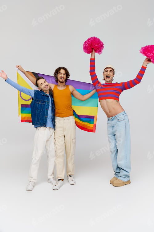 Preview: positive appealing gay men in vibrant clothes posing with rainbow flag and pom poms on gray backdrop