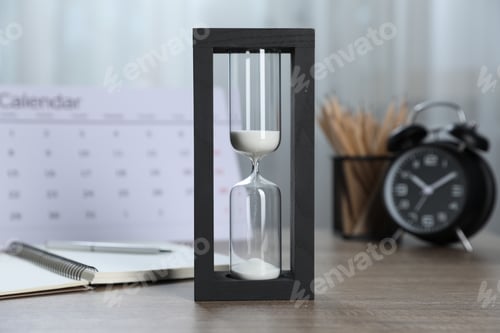 Preview: Deadline. Hourglass with flowing sand, stationery, calendar and alarm clock on wooden table indoors