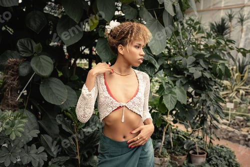 Preview: Stylish young african american woman in summer outfit looking away while standing near green plants