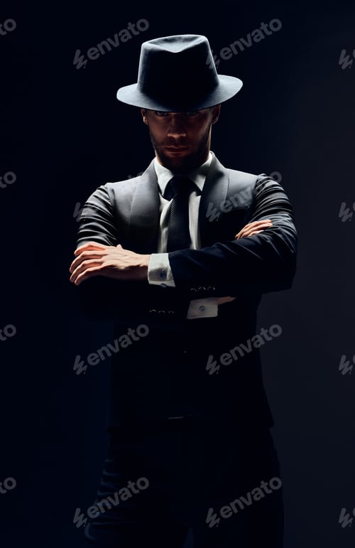 Preview: Handsome confident man in black suit and hat with arms crossed on dark background