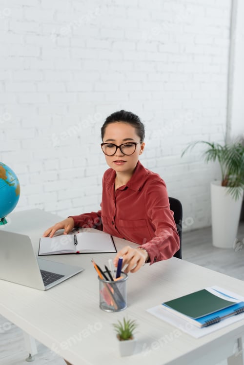 Preview: young teacher in eyeglasses reaching stationery near laptop on desk