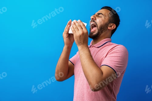 Preview: Young man in pink shirt with allergy or cold, blowing his nose in a tissue against blue background