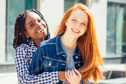 Preview: cheerful afro and european lesbian women laughing and looking at camera at summer downtown city