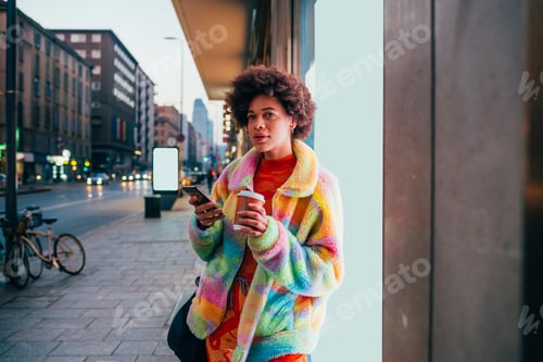 Preview: Young black woman outdoors at night using smartphone drinking from reusable cup