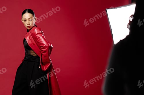 Preview: Fashion asian woman in red coat and black pants posing for photographer in front of red background