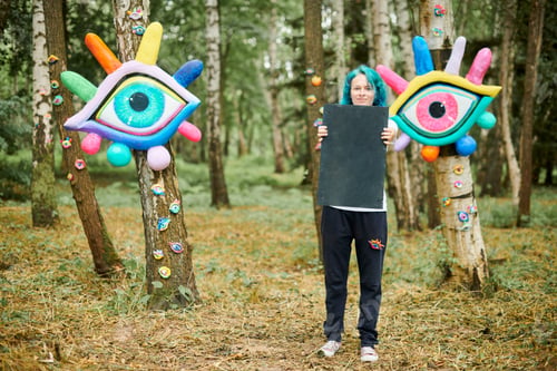 Preview: Funny young adult woman with dyed turquoise hair in white T shirt near big eye art installation